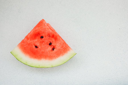 A slice of watermelon rests on a plain surface with a light color. The pink flesh and green rind are visible. It shows a casual scene where fruit is prepared for eating.の写真素材