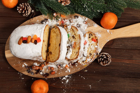 A wooden board holds slices of a stollen cake covered in powdered sugar. The cake is adorned with dried fruits nuts and placed beside fresh mandarins and pine cones during the holiday period.の写真素材