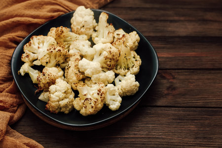 Oven roasted cauliflower pieces are arranged neatly on a black plate. A brown cloth is nearby on a wooden table surface. This setting shows a simple food preparation scene.の写真素材