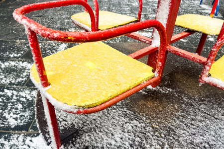 Bright yellow seats on playground equipment are covered in snow during a cold winter day. The surrounding ground is also blanketed in white highlighting the quiet and chilly atmosphere.の写真素材