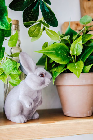 A rabbit statue sits beside a potted plant on a wooden shelf. The plant has green leaves. The...