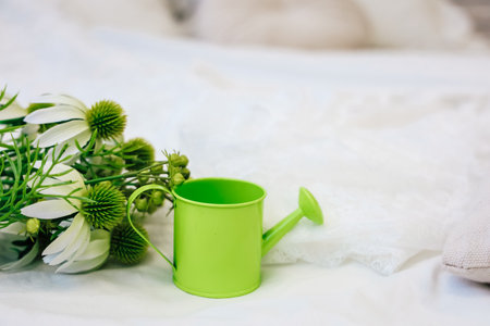 A small green watering can sits next to white flowers on a bed. The bedding is light and soft. The flowers add a touch of nature to the indoor space. This setup is inviting and pleasant.の写真素材