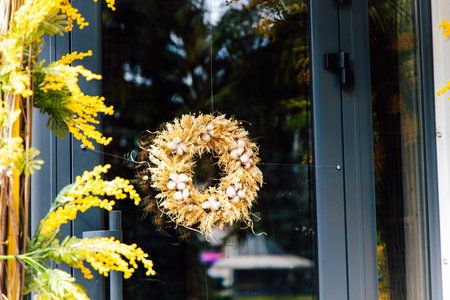 A round wreath made from straw and small flowers is placed on a door. Yellow plants are seen on either side of the door. The door is glass and reflects a garden in the background.の写真素材