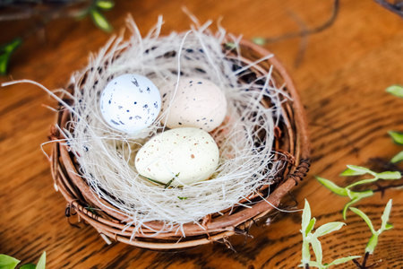 Three eggs sit in a nest made of twigs and grass on a wooden table. The eggs have different colors...