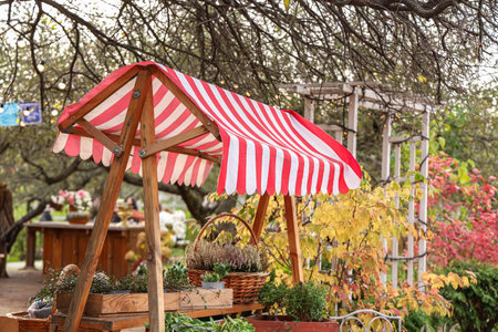 A garden market stand with a red and white striped canopy is surrounded by plants and trees during autumn. People walk by and enjoy the fall colors in the park.