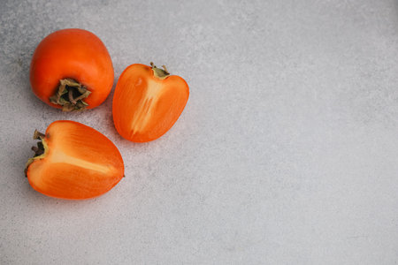 Persimmons are arranged on a flat gray surface. Two fruits are halved to reveal their bright orange flesh. One whole persimmon rests beside the sliced pieces in a simple display.