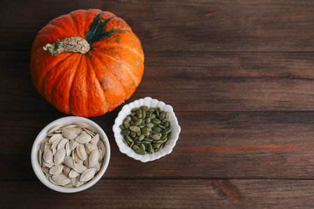 A pumpkin rests on a wooden table beside two bowls filled with pumpkin seeds and pepitas. This scene captures the essence of fall preparations and cooking.