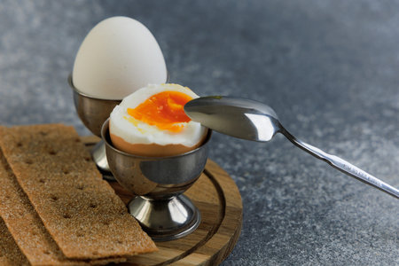 A boiled egg sits in a metal holder on a wooden plate. A spoon is used to scoop out the yellow yolk. Crackers are placed beside the egg.