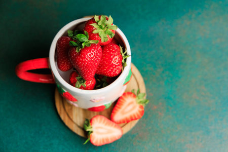 A white cup filled with fresh strawberries sits on a wooden board. Sliced strawberries lay nearby. The background is green creating a vibrant scene.