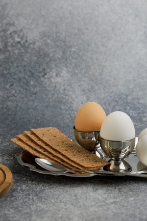 An arrangement shows two boiled eggs in silver holders next to sliced crispbread on a silver tray. A wooden board is nearby on a grey surface.