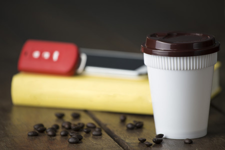 Hot coffee white plastic cup with brown cap and car key and mobile and blank yellow book cover on old wooden table with low lightの写真素材