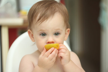 cute blond child sits in a children's chair at the kitchen and eat a juicy peach. Little girl with a piercing lookの写真素材