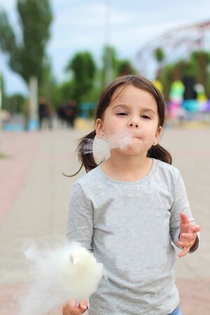 Little funny white girl with tails with narrow eyes holds cotton candy with his lips while walking outdoorsの写真素材