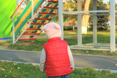 A small child in a fluffy pink hat and red vest is running in the autumn park. Beautiful fall sunny day outdoors.の写真素材