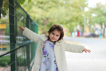 A little girl with long dark hair in a fluffy white coat stands in front of a metal fence. Blurred back street backgroundの写真素材