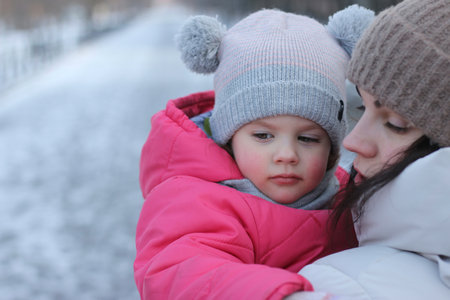 Little girl in a knitted hat with buboes in the arms of a smiling mother on the street in winterの写真素材