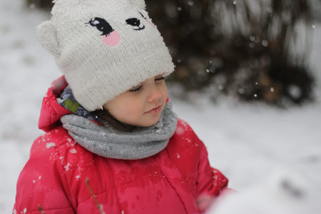 A small beautiful cute child in warm clothes and a knitted hat with a snowflake stuck to his lip stands with closed eyes and a satisfied faceの写真素材