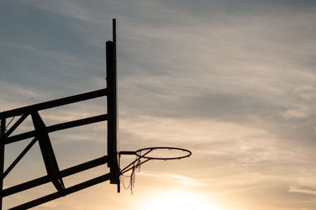 An underexposed image of a basketball backboard with a hoop against a gloomy sunset skyの写真素材