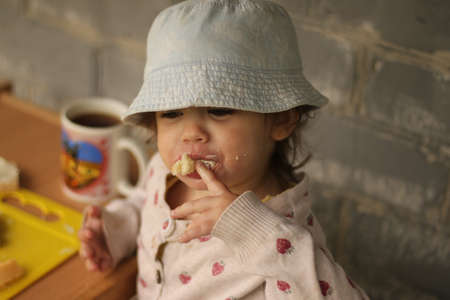 A funny child in a Panama hat and a sweater eats at the table against the background of a brick wallの写真素材