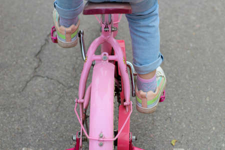 Image of a pink bicycle on which a child in jeans and sneakers rides on the asphaltの写真素材