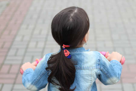 Centered image of a little white girl with dark ponytail hair riding a bicycle in a denim jacket on a blurry brick road backgroundの写真素材