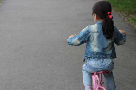 A little girl in a denim suit rides a pink bicycle on the asphalt on the right side. Space for text on the leftの写真素材