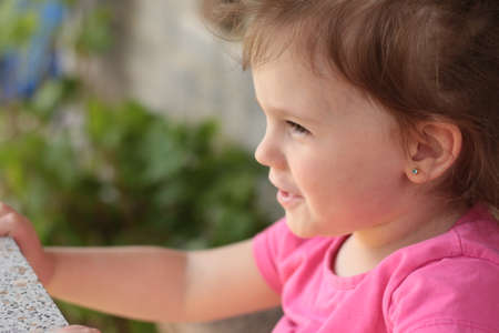 Beautiful little smiling red-haired girl in a pink t-shirt sits at a table outdoorsの写真素材
