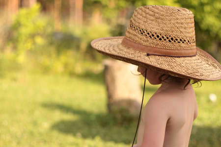 Photo of a sad child in a straw farm hat standing from the side against the backdrop of a blurred vineyard landscapeの写真素材