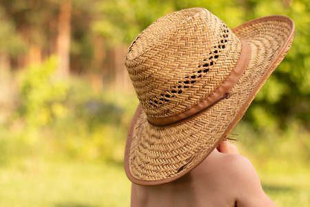 A small lovely child in a straw farm hat looks into the distance with his back to the camera. Wonderful childhood moments on the farmの写真素材