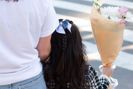 A little girl with long dark hair walks through the pedestrian crossing holding her mother's hand. Child goes to school with a bouquet of flowersの写真素材
