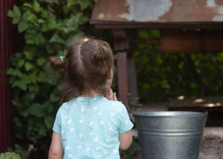 little smiling country girl looks thoughtfully at the metal bucket on the wellの写真素材