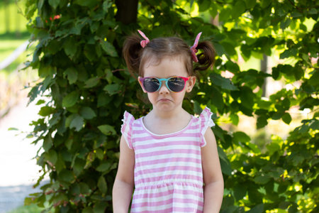 Little girl in a striped dress and sunglasses sadly looks at the camera against a blurred deciduous...