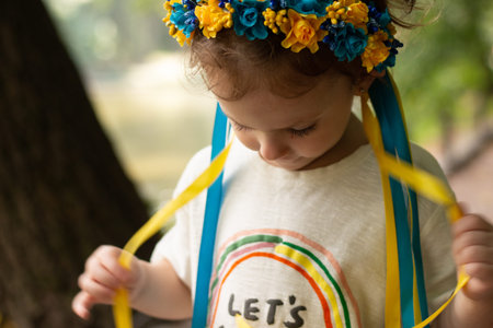 Sad girl with Ukrainian folk decoration on her head. Little kid touching wreath ribbons while...