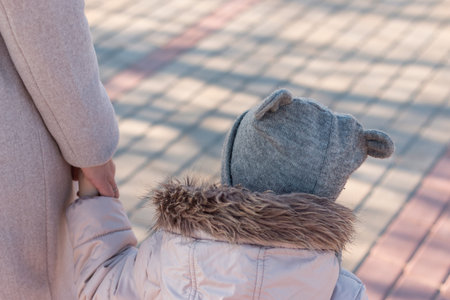A small child in a funny hat and jacket with a fluffy hood holds a parent's hand while walking...