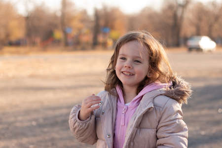 Beautiful red-haired little girl in a jacket with a fluffy hood smiles on the background of an autumn streetの写真素材