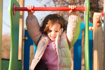 Small child with long red hair against the background of a blue sky in warm clothes on the playgroundの写真素材