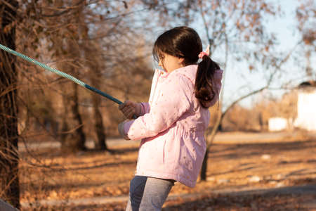 A small child with long hair gathered in a ponytail against the background of a blue sky in warm clothes on the playgroundの写真素材