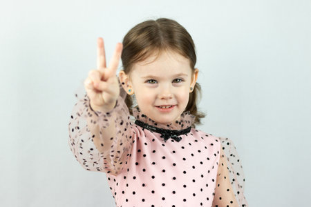 Smiling little girl with two ponytails in a dress with polka dots on a white background shows two fingers raising her hand forward. Studio photoの写真素材