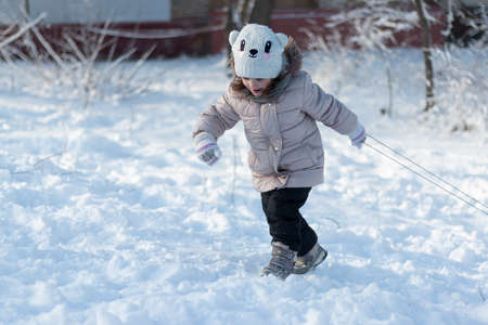 A small child in warm clothes pulls the sled uphill with difficulty. Outdoor photo shoot on a sunny winter dayの写真素材