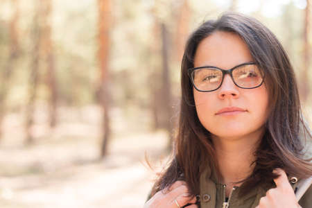Close-up portrait of a girl with dark long hair in glasses standing with a backpack against the background of blurry trees in the forestの写真素材