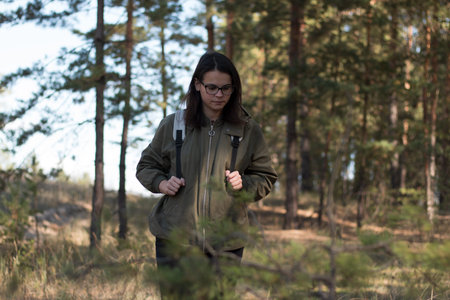 cute girl with dark long hair in glasses looks down standing in the forest with a backpackの写真素材