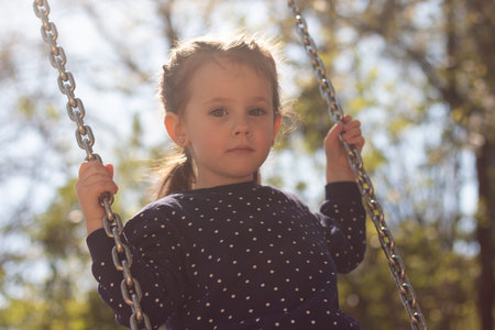 Little serious girl with braided pigtails rides on a swing in the parkの写真素材