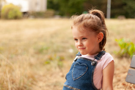 A beautiful blonde little girl with a tail sits on a bench in the park in a denim overallsの写真素材