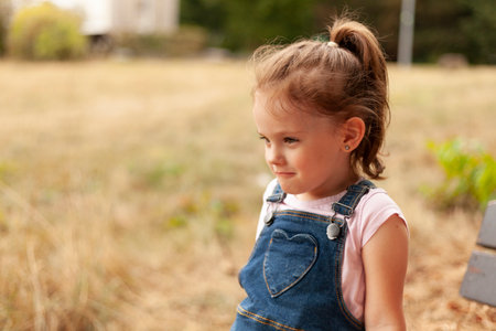 A beautiful blonde little girl with a tail sits on a bench in the park in a denim overallsの写真素材