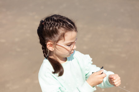 A little girl in glasses is winding her wind kite while standing on a sandy beach.