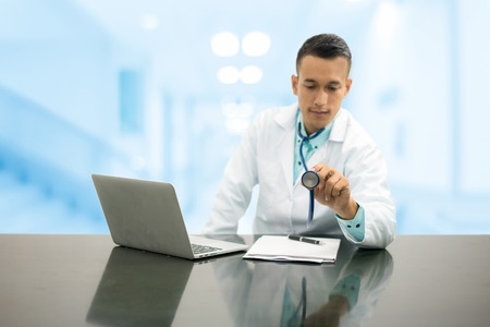 Male doctor pointing stethoscope at copy space, sitting on office desk with computer and paperwork against hospital background. Medical and healthcare concept.の写真素材