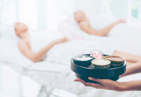 Massage therapist holding spa treatment set and hot scrub lotion with woman lying on spa bed prepared for spa massage in background. Luxury wellness, stress relief and rejuvenation concept.の写真素材
