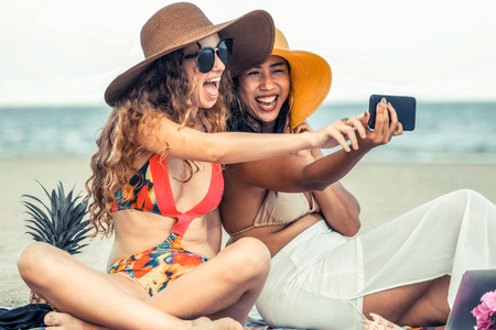Happy women in bikinis taking selfie photograph from mobile phone together on tropical sand beach in summer vacation. Travel lifestyle.の写真素材