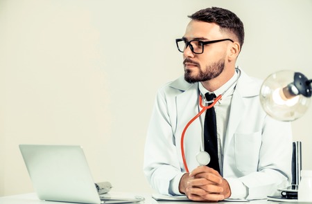 Happy male doctor at office desk in hospital working on laptop computer. Medical healthcare concept.の写真素材
