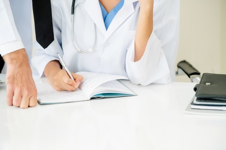 Doctor at hospital office writes notes on patients report while having conversation with another doctor that standing beside her.の写真素材